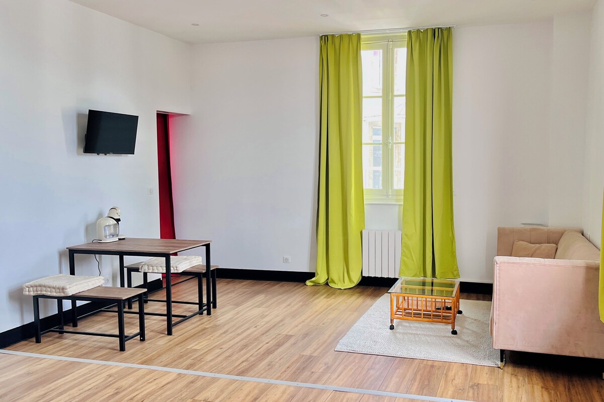 Quarto com piso de madeira, uma mesa com cadeira, um pequeno sofá, um radiador e uma janela com cortinas.