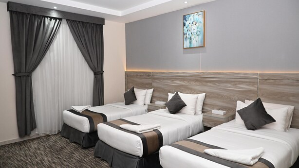 A hotel room with three beds, a large window with curtains, a painting on the wall, and a wooden headboard. - Jawharat Alalia Hotel Apartments (Madinah)