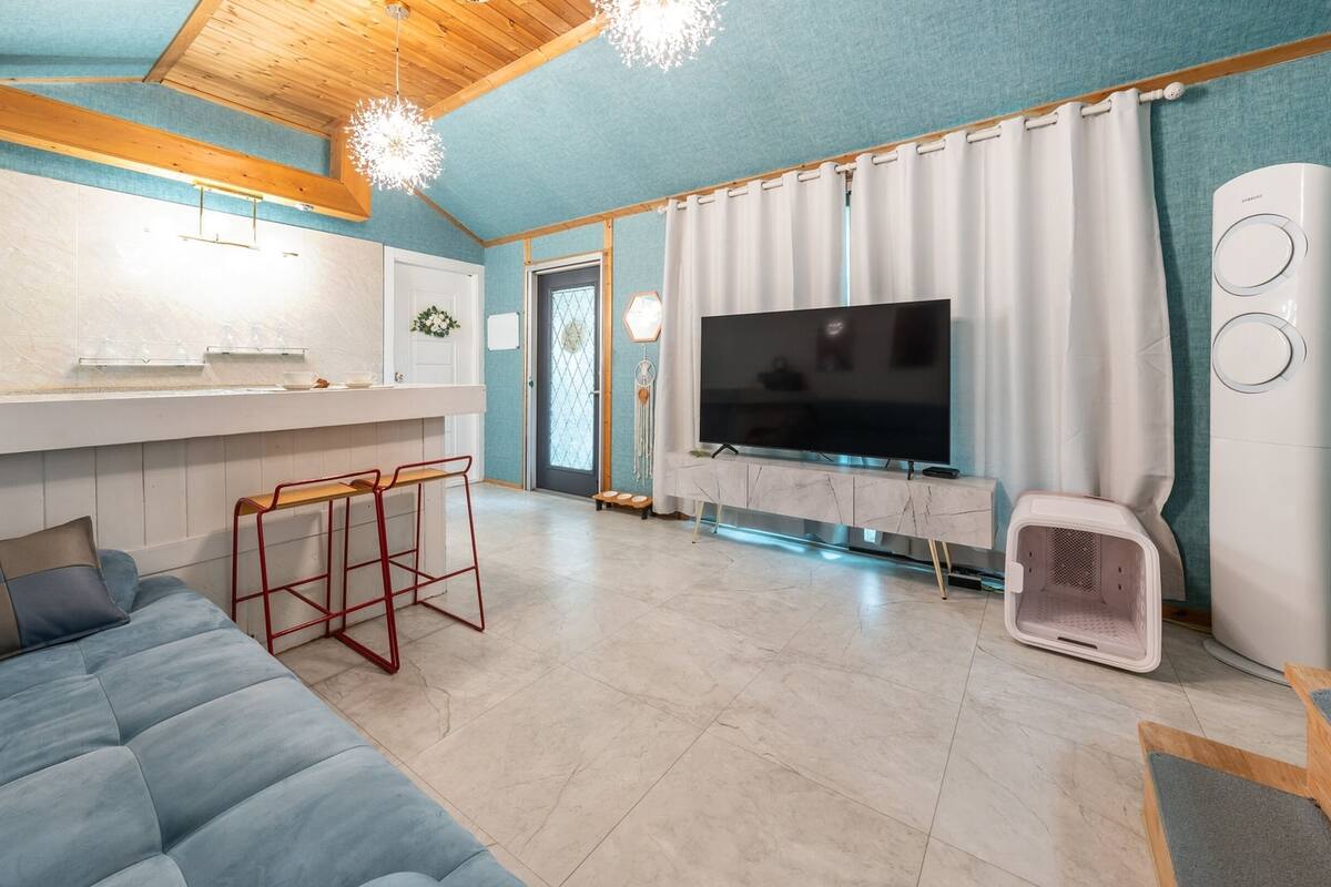 Una sala moderna con sofá, televisión, transportín para mascotas y una cocina con mostrador y taburetes.