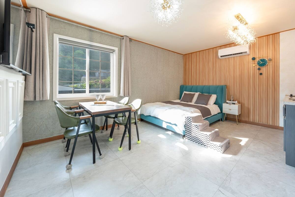 Un dormitorio moderno con cama, escritorio y silla. Hay una ventana, un aire acondicionado y un reloj en la pared.