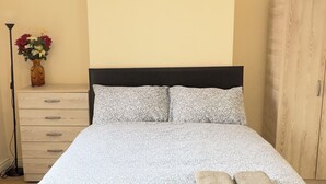 Ein ordentlich bezogenes Bett mit einem blumigen Überwurf, eine Uhr an der Wand und ein Holzschrank zur Rechten.