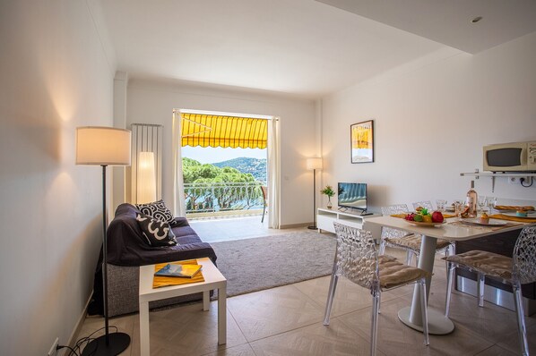 A modern living room with a sofa, a dining table with chairs, a TV, and a balcony with a yellow awning.