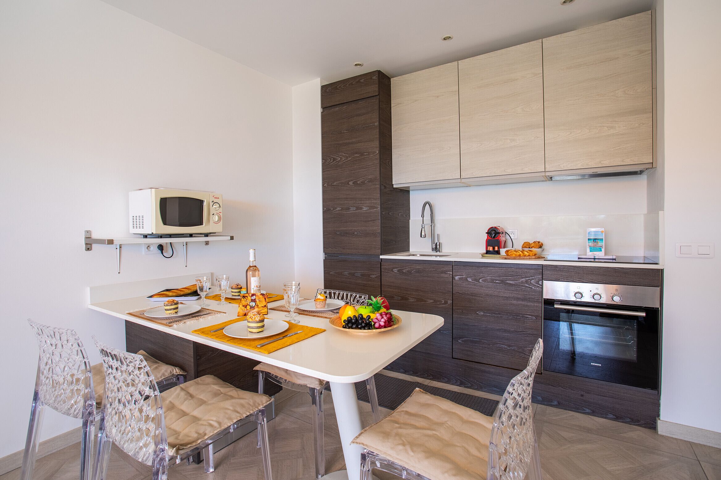 A modern kitchen with a microwave, oven, and wooden cabinets. There is a dining table with chairs and a fruit bowl.