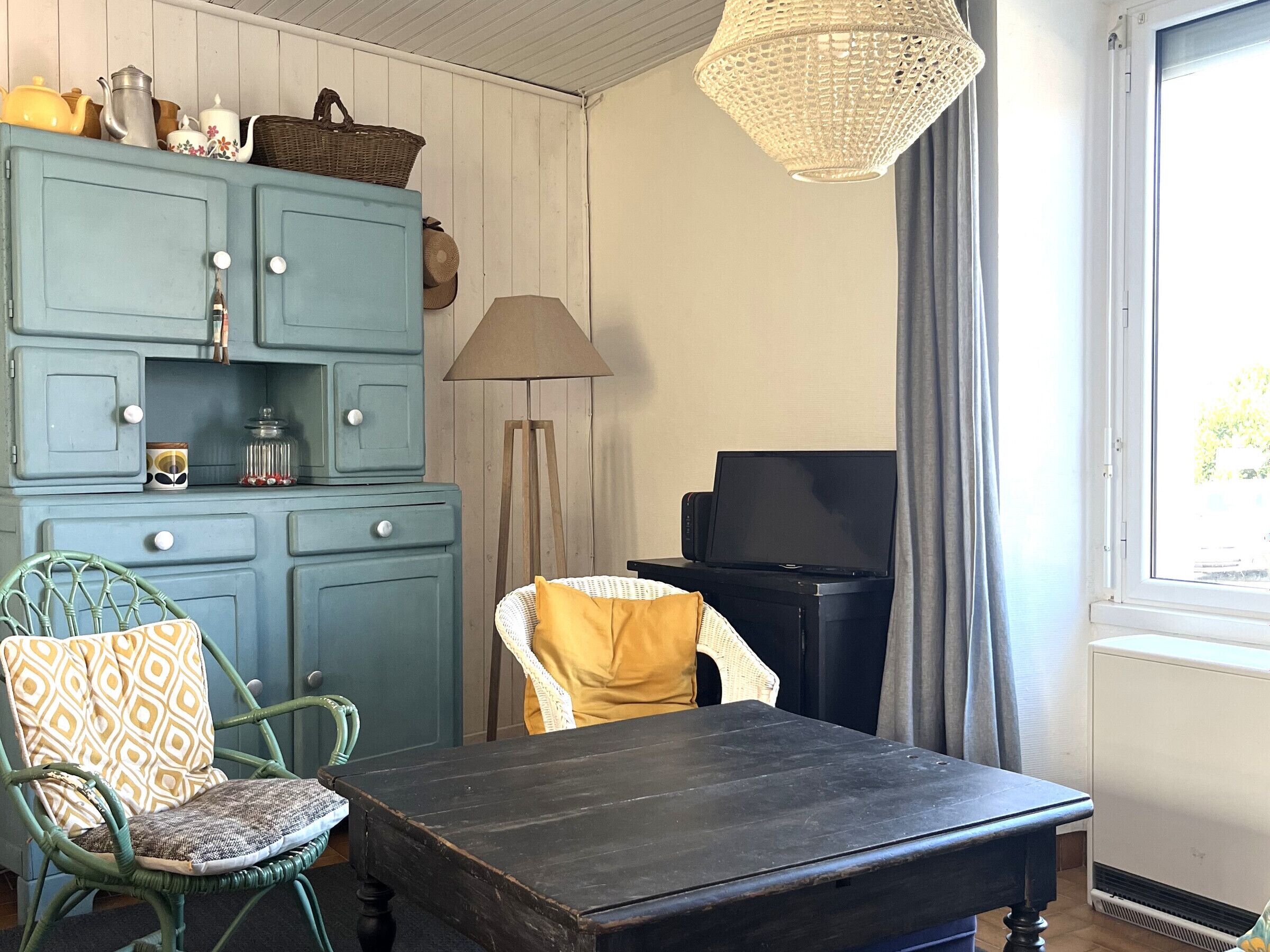 A room with a blue cabinet, a wicker chair, a table, a lamp, and a television.