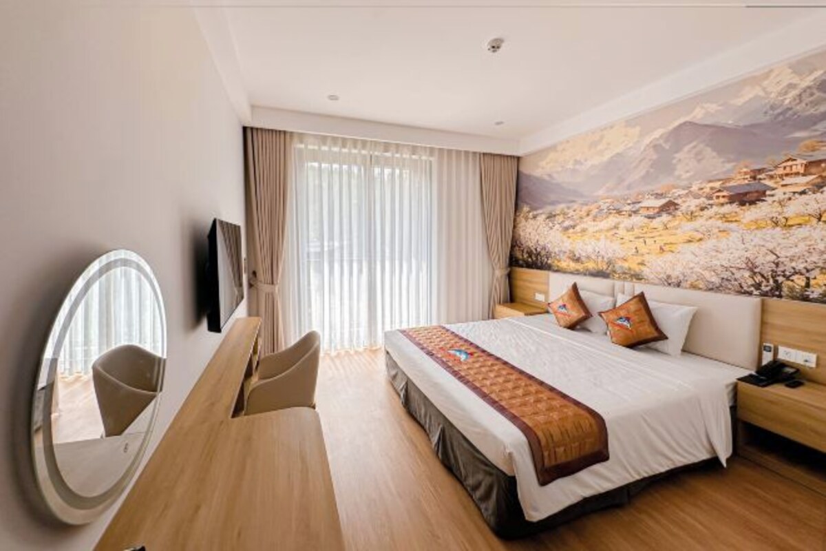 Habitación de hotel con una cama grande, un escritorio, una silla, un espejo redondo y un cuadro de un paisaje montañoso.