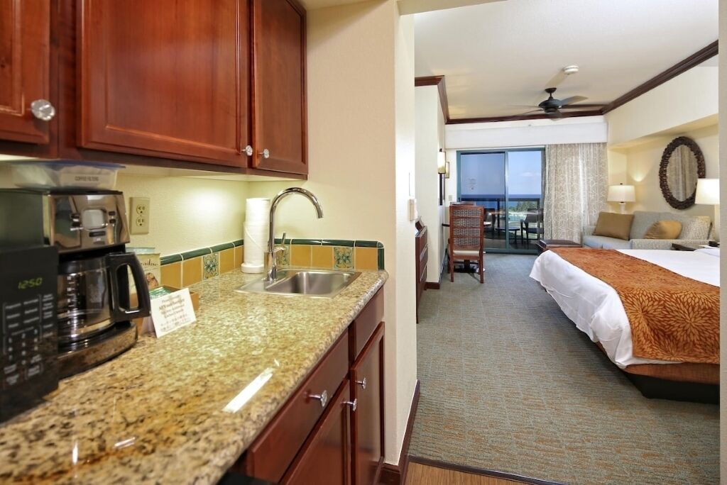 A hotel room with a kitchenette, a bed, a view of the balcony, and a large window.