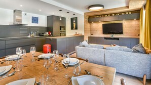 A modern kitchen with a dining area, a grey sofa, and a flat-screen TV mounted on the wall.