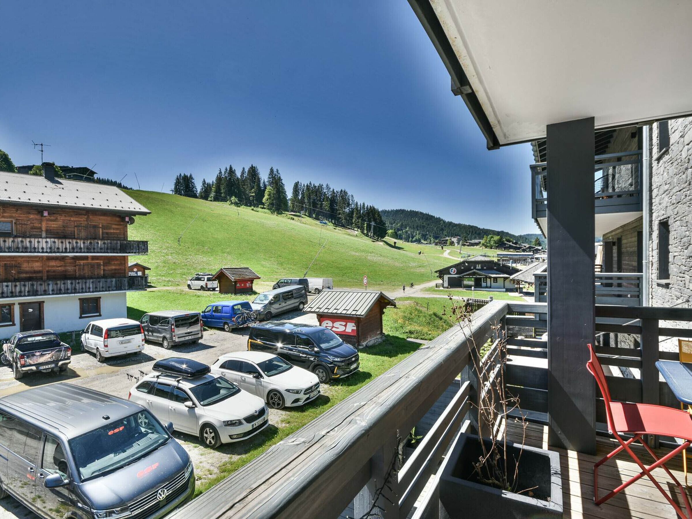 Ein Parkplatz mit verschiedenen Fahrzeugen, ein Gebäude mit Holzfassade und Blick auf einen grünen Hügel.