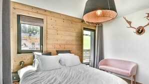 A bedroom with a large bed, a wooden headboard, a window with a view, a pink chair, and a wall-mounted deer head decoration.