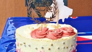 A cake with a white and pink gradient design, decorated with roses and gold beads, placed on a white plate on a blue tablecloth.