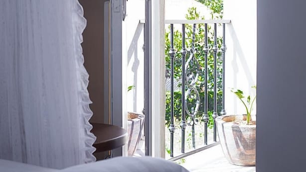 A bedroom with a large window, a white bed, and a view of a balcony with a railing and potted plants.