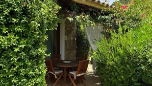 A patio with a round table and chairs, surrounded by lush greenery and a stone wall. - Villa daisy, Chia (Sardegna)