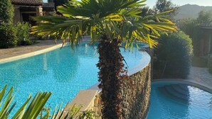 A pool with a stone wall and a palm tree, surrounded by greenery and a building in the background. - Villa daisy, Chia (Sardegna)