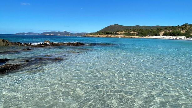 A coastal landscape with clear turquoise waters, rocky shoreline, and a distant beach with people. - Villa daisy, Chia (Sardegna)