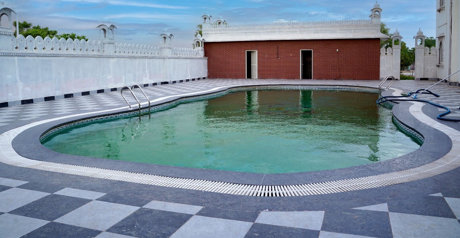 A swimming pool with a checkered floor, surrounded by a white wall with decorative elements and a red brick building in the background.
