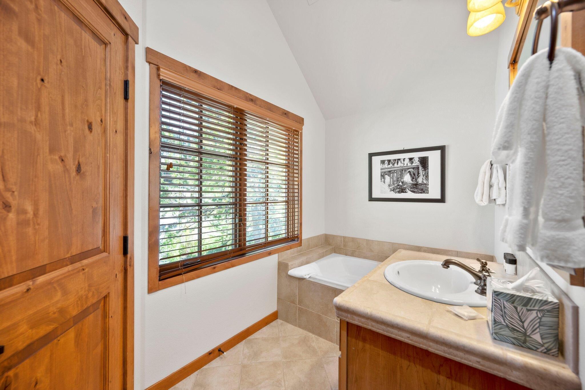 A bathroom with a wooden door, a window with blinds, a sink with a mirror, and a towel rack.
