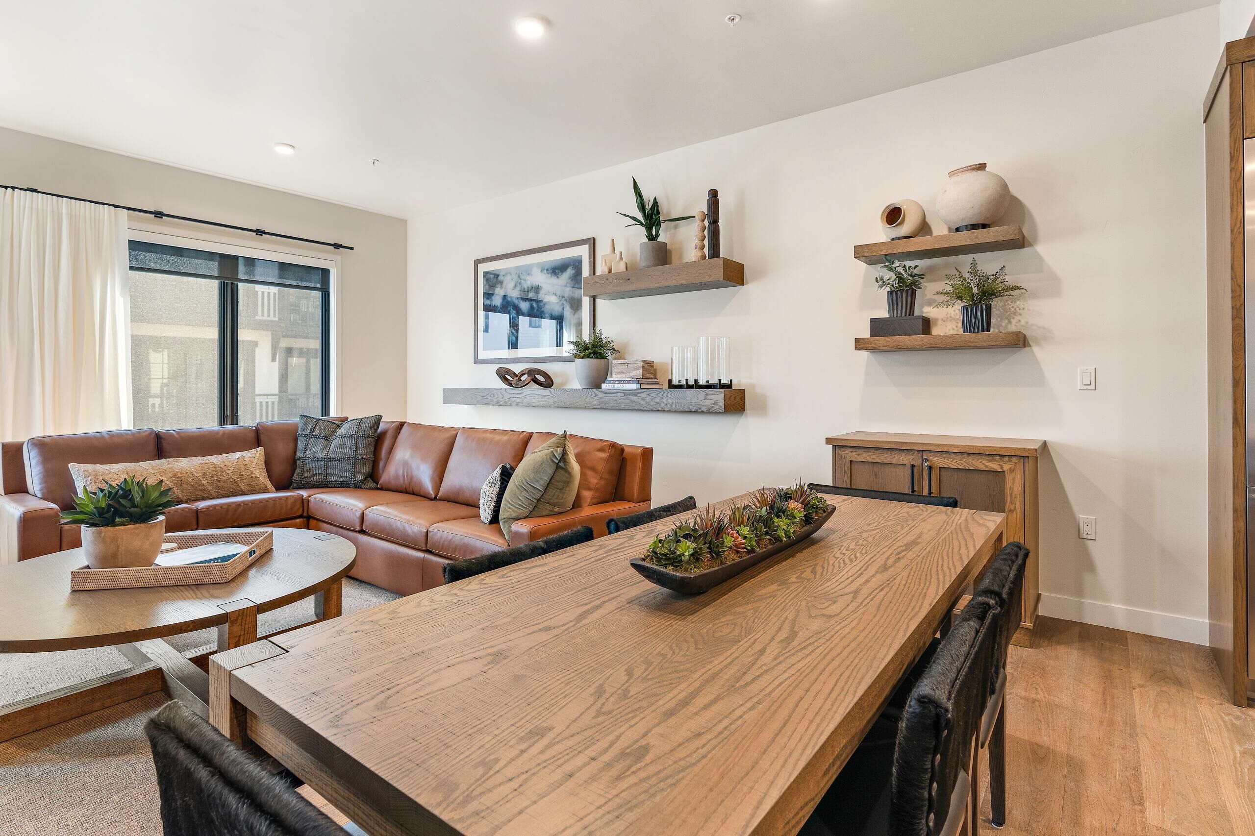 A modern living room with a wooden dining table, leather sofa, and built-in shelves with decorative items.