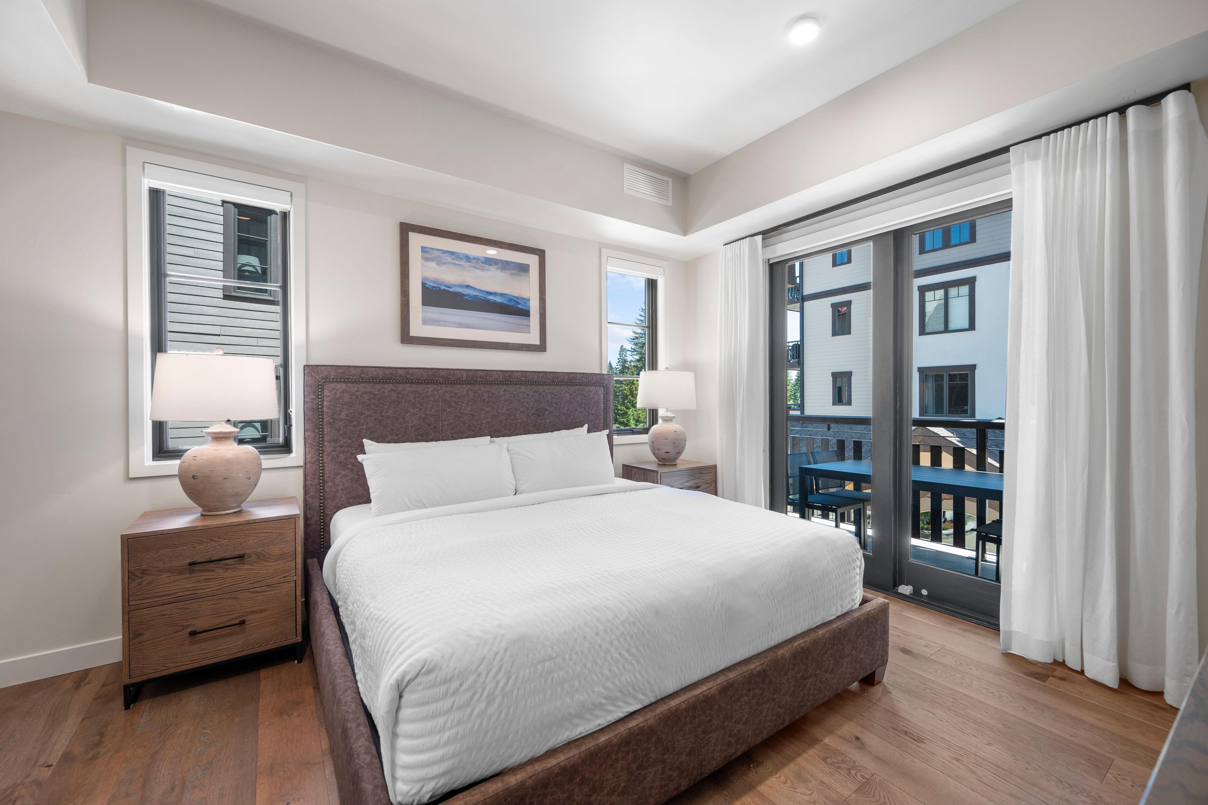 A modern bedroom with a large bed, two bedside tables, a framed picture on the wall, and a view of the outside through a sliding glass door.