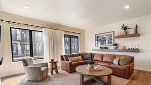 Una sala de estar moderna con un sofá de cuero marrón, un sillón gris y una mesa de centro de madera. Hay un ventanal con cortinas, una mesita auxiliar y una estantería con objetos decorativos.