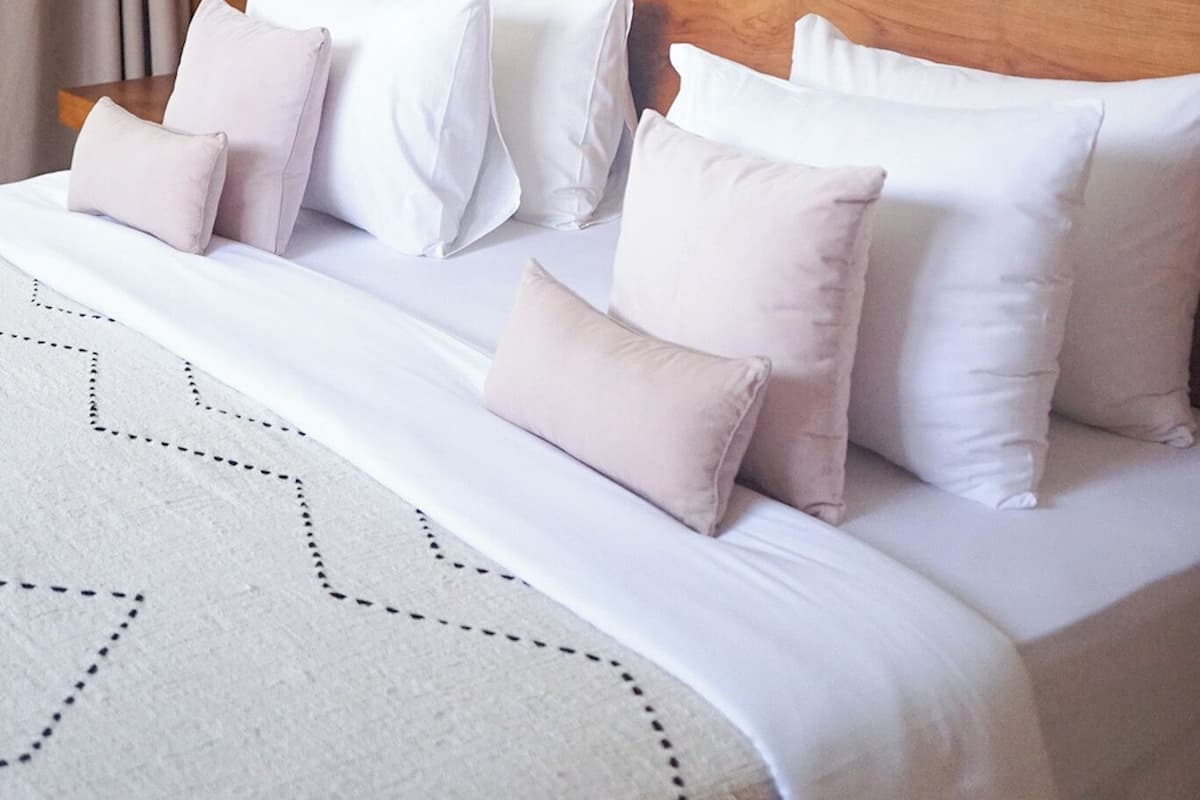 A neatly made bed with white and light pink pillows, a woven wall art piece, and a patterned rug.