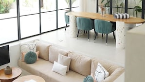 A spacious living room with a large window, a ceiling fan, a wooden dining table, and a sofa with cushions. - Arrova Bali Berawa, Canggu by Sandara (Jimbaran)
