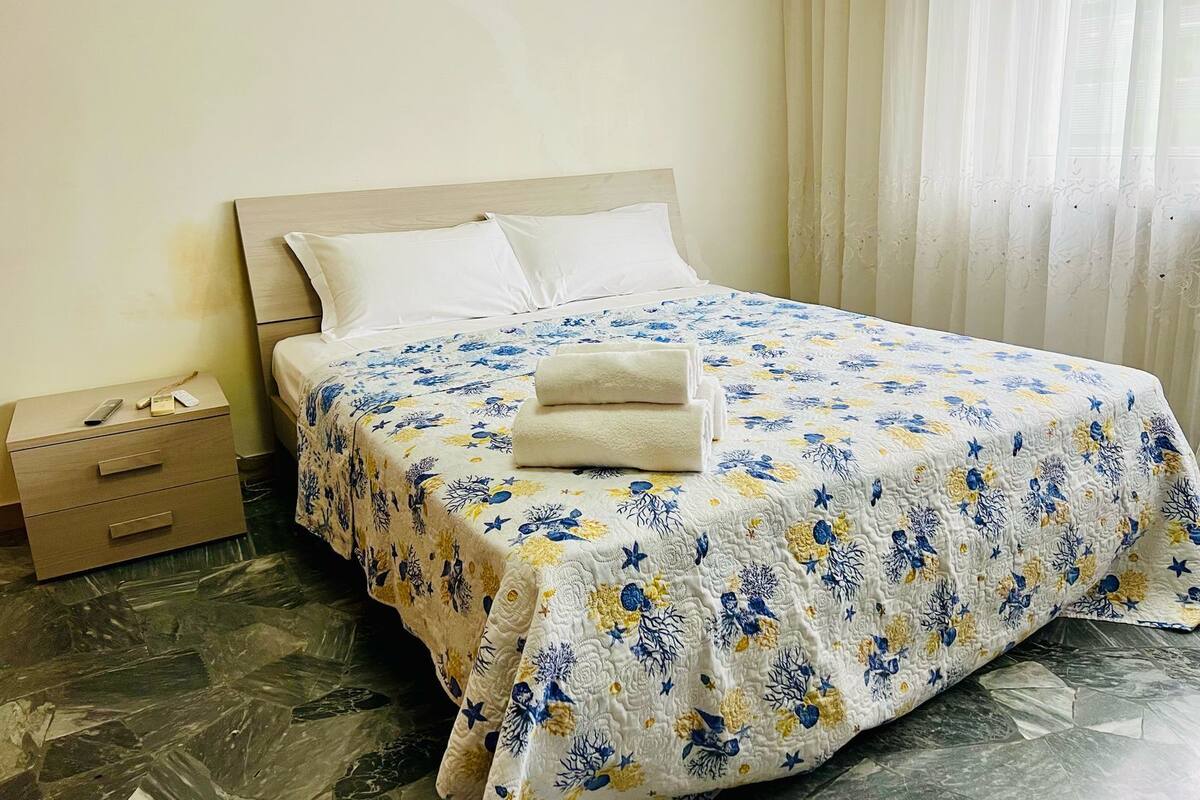A neatly made bed with a floral bedspread, a bedside table with a remote control, and a drawer unit.