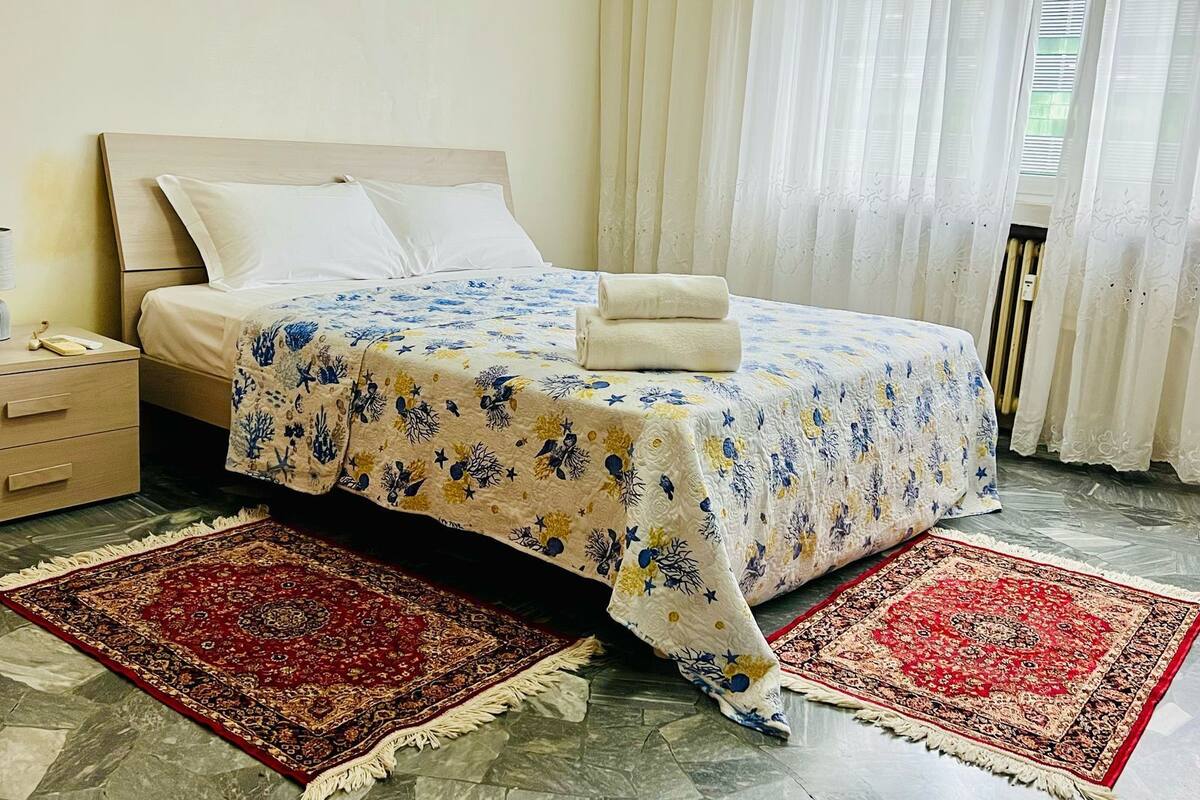 A neatly made bed with a floral bedspread, two red and beige area rugs, a wooden nightstand with a lamp, and a window with sheer curtains.