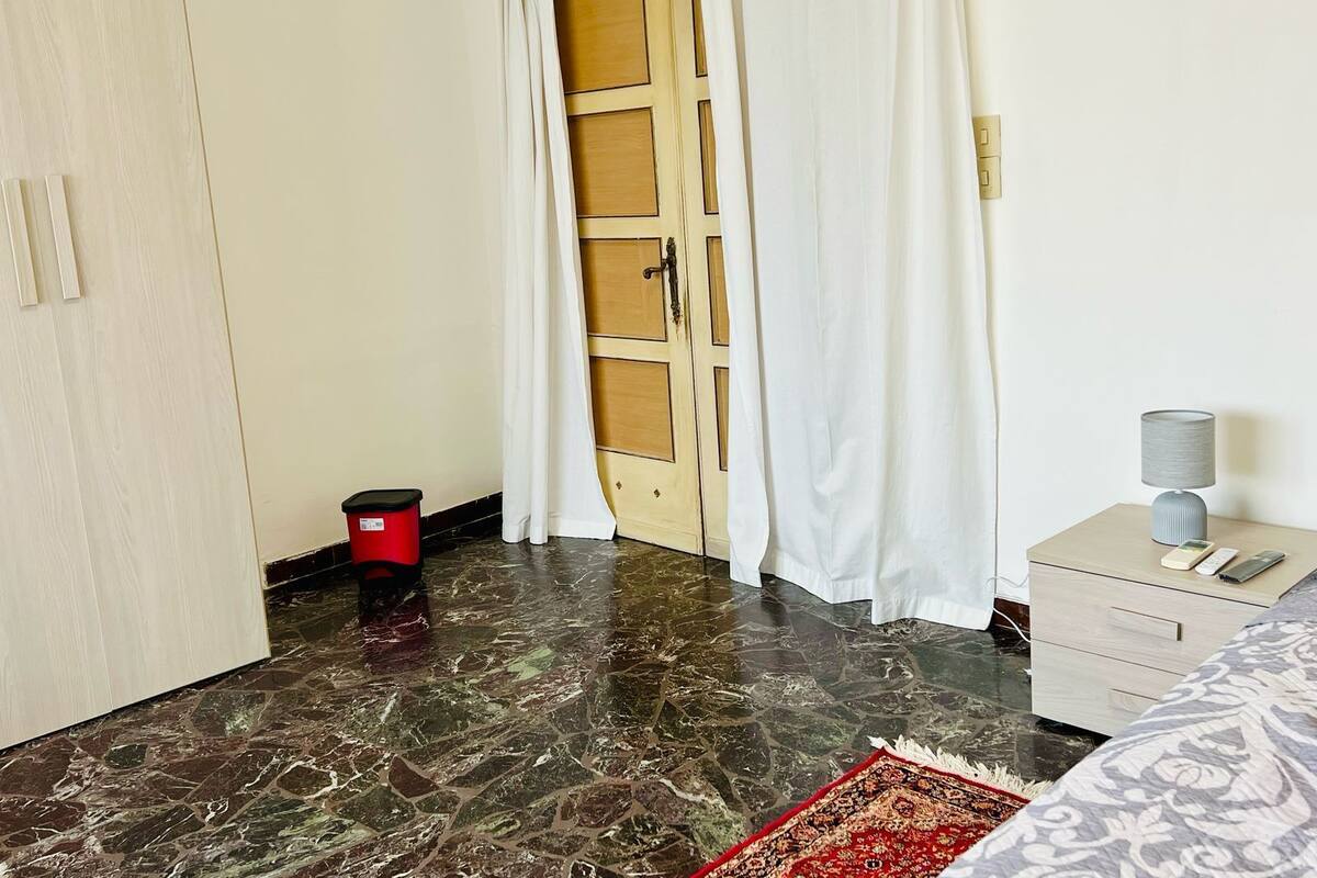 A hotel room with a marble floor, a wooden door, a small trash bin, a bedside table with a lamp, and a patterned bedspread.