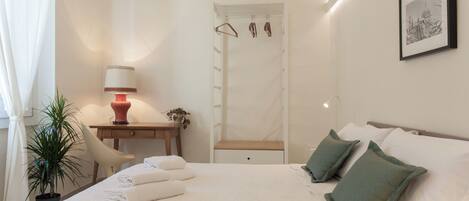 Habitación de hotel con cama, almohadas, escritorio con lámpara, una silla, planta y puerta con perchero.