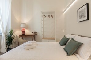 Ein Hotelzimmer mit Bett, Kissen, Schreibtisch mit Lampe, Stuhl, Pflanze und Tür mit Kleiderhaken.