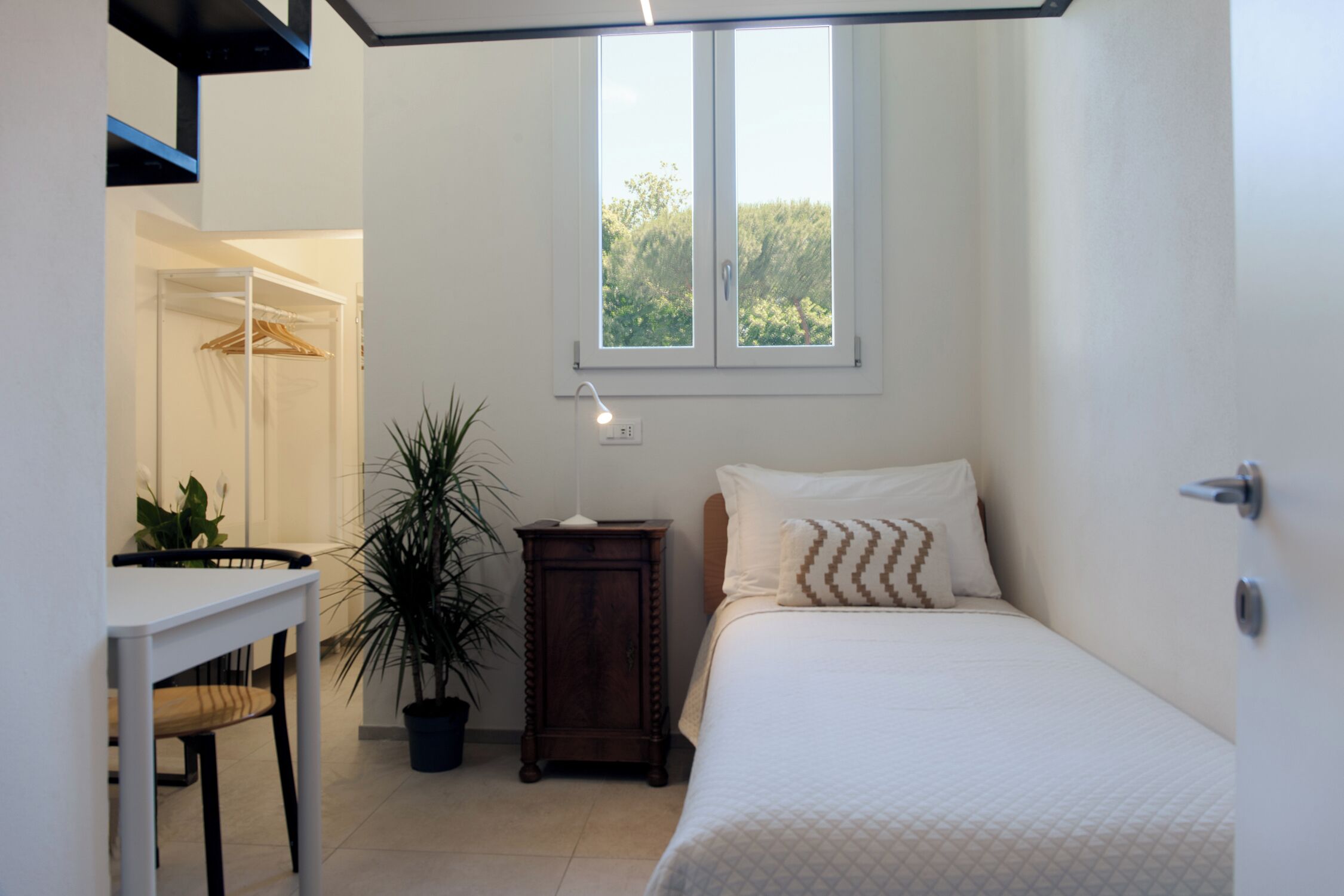 Ein Schlafzimmer mit Bett, Nachttisch, Blumentopf und Fenster mit Blick auf Grünflächen.