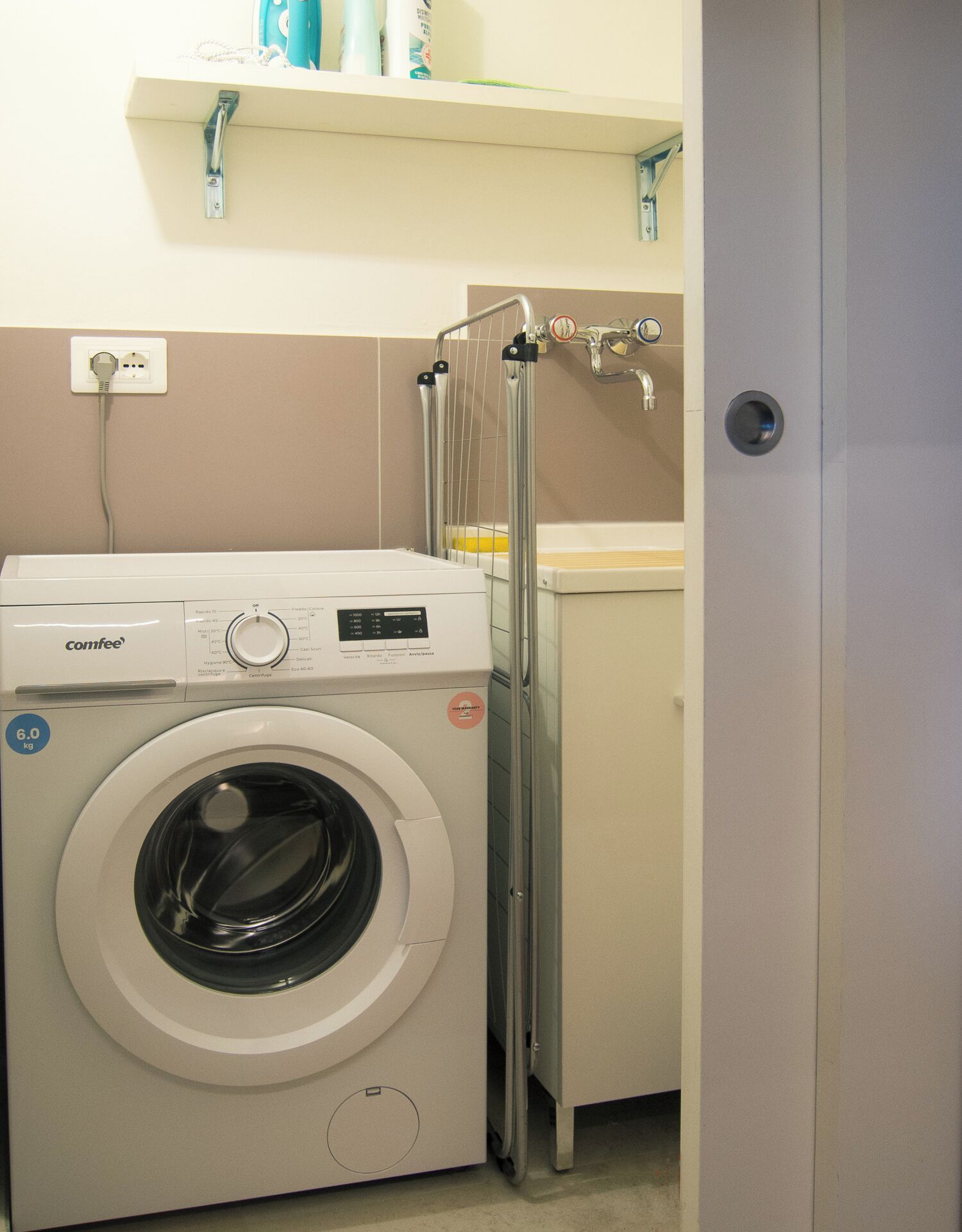 A compact, modern laundry room with a front-loading washing machine, a drying rack, and a small shelf above the machine.