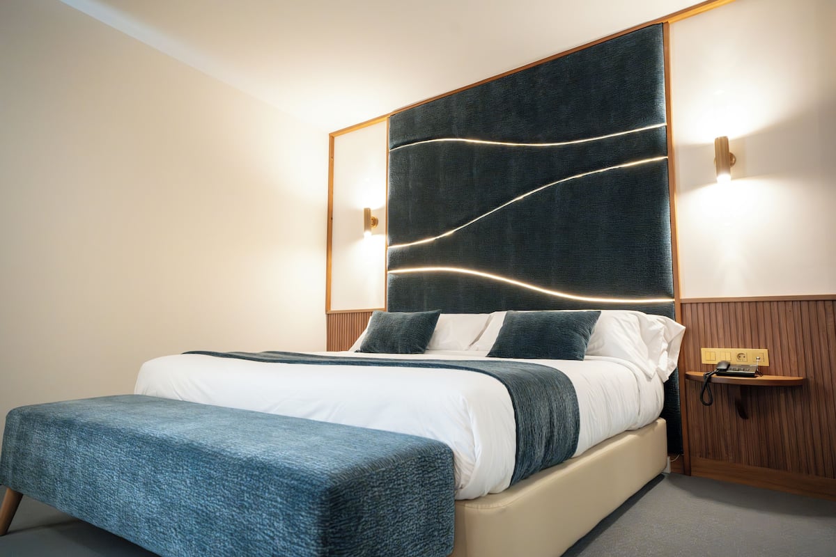 Ein modernes Hotelzimmer mit einem großen Bett, einem Nachttisch mit Telefon und einem Kopfteil mit integrierter Beleuchtung.