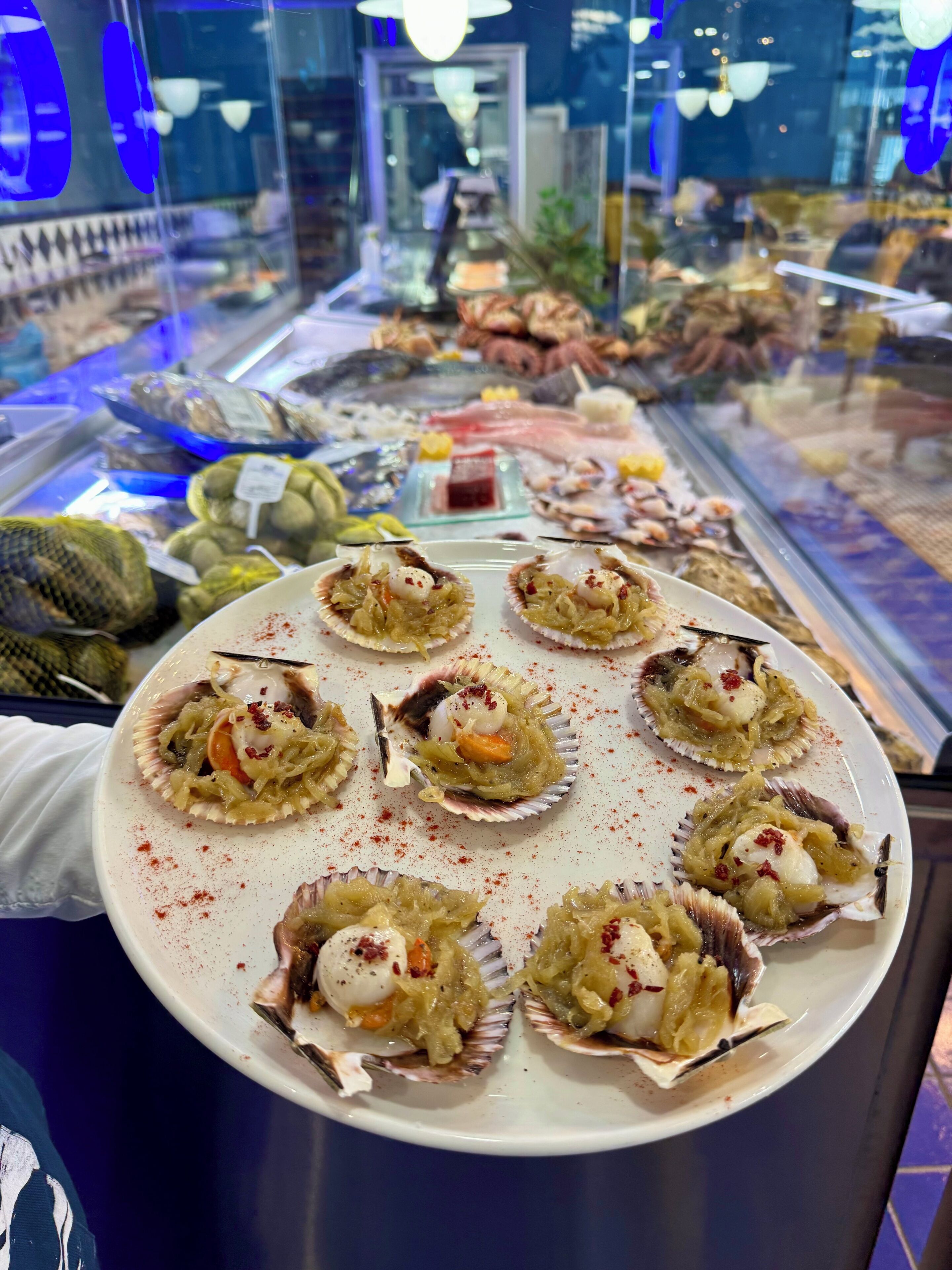Eine Platte Jakobsmuscheln mit Garnitur vor einer Fischvitrine.