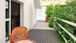 Een balkon met een houten vloer, een rieten stoel en een kleine tafel. Er is een witte balustrade en uitzicht op een gebouw met een pannendak.