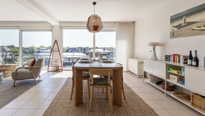 A modern living room with a dining table, chairs, a hanging light fixture, a bookshelf, and a large window offering a city view. - 3 bedroom accommodation in Sint-Idesbald (Sint-Idesbald)