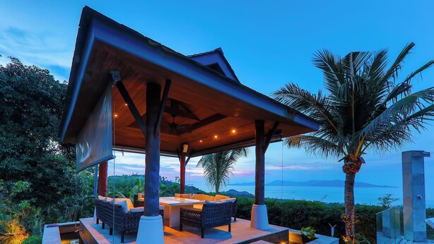 A covered outdoor seating area with a pool, surrounded by lush greenery and overlooking a body of water.