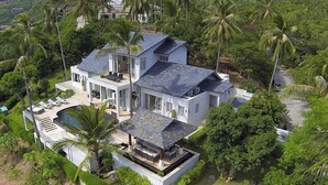 A modern two-story house with a swimming pool, surrounded by lush greenery and palm trees.