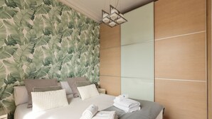 A neatly made bed with pillows and towels, a wooden wardrobe, and a wall with a leafy pattern. - Enjoy Apartments Donna (Barcelona)