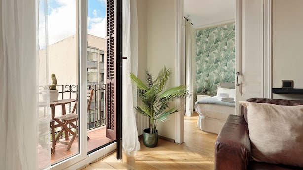 A room with a wooden floor, a brown leather sofa, a potted plant, and a view of a balcony with a table and chairs. - Enjoy Apartments Donna (Barcelona)