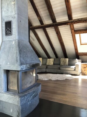 Ein Kamin aus Stein mit Lüftung, eine Holzbalkendecke, ein graues Sofa mit Kissen und ein weißer Teppich auf einem Holzboden.