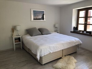 Ein ordentlich bezogenes Bett mit weißer Tagesdecke und zwei Kissen, ein Nachttisch mit Lampe, ein gerahmtes Bild an der Wand und ein Fenster mit Holzrahmen.