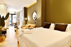 A modern hotel room with a large bed, a seating area with a sofa and chairs, a dining table, and a large potted plant.