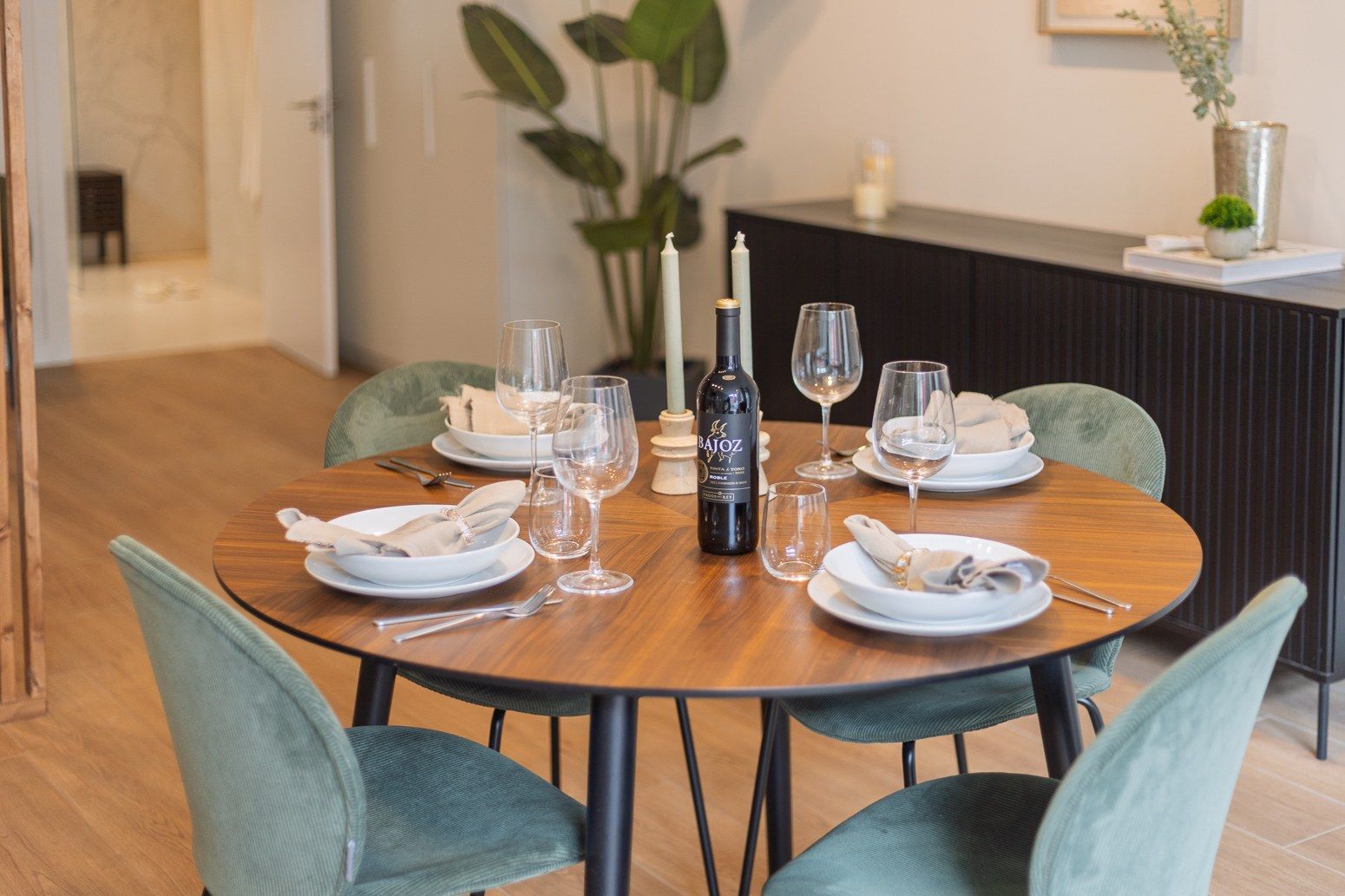 Una mesa de comedor dispuesta con platos, cubiertos, copas y una botella de vino, rodeada de sillas.