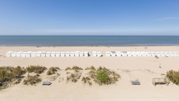 A beach with rows of white beach huts and a sandy foreground with some vegetation.