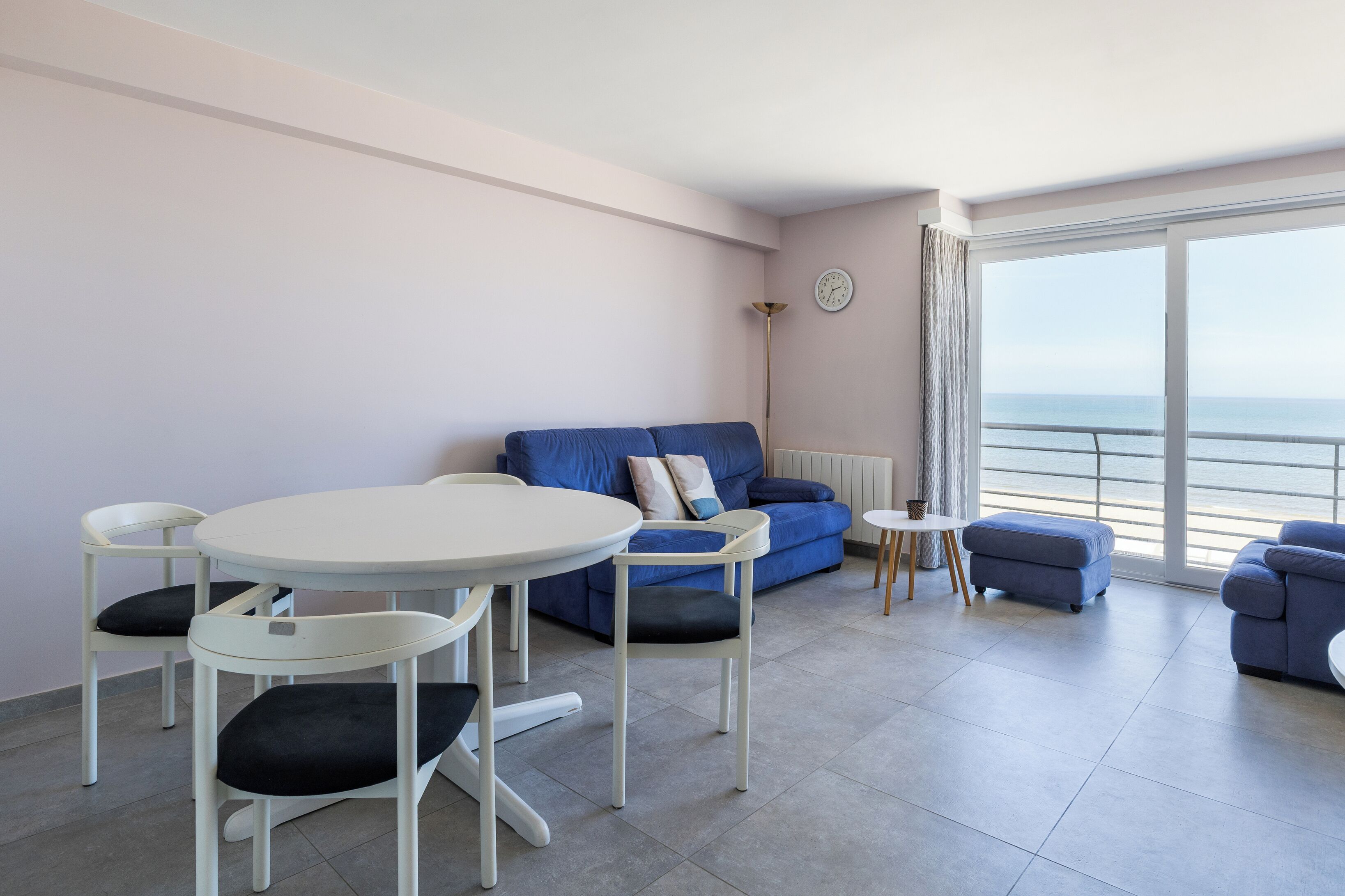 A modern living room with a blue sofa, white chairs, a small round table, and a view of the sea through large windows.