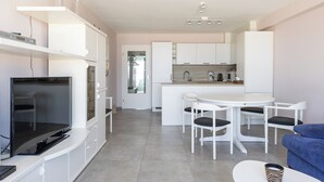 A modern kitchen with white cabinets, a dining area with a white table and chairs, and a flat-screen TV on a white entertainment unit.
