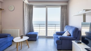 A modern living room with a sofa, a small table, a TV, and a view of the sea through large windows.