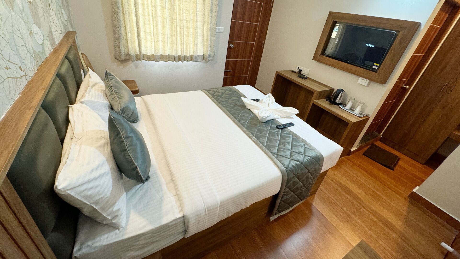 A neatly made bed with pillows and a blanket, a bedside table with a remote control, a television mounted on the wall, and a wooden floor.