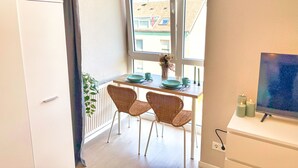 Un petit balcon avec une table dressée pour deux personnes, une chaise en osier, une plante en pot et une vue sur les bâtiments environnants.